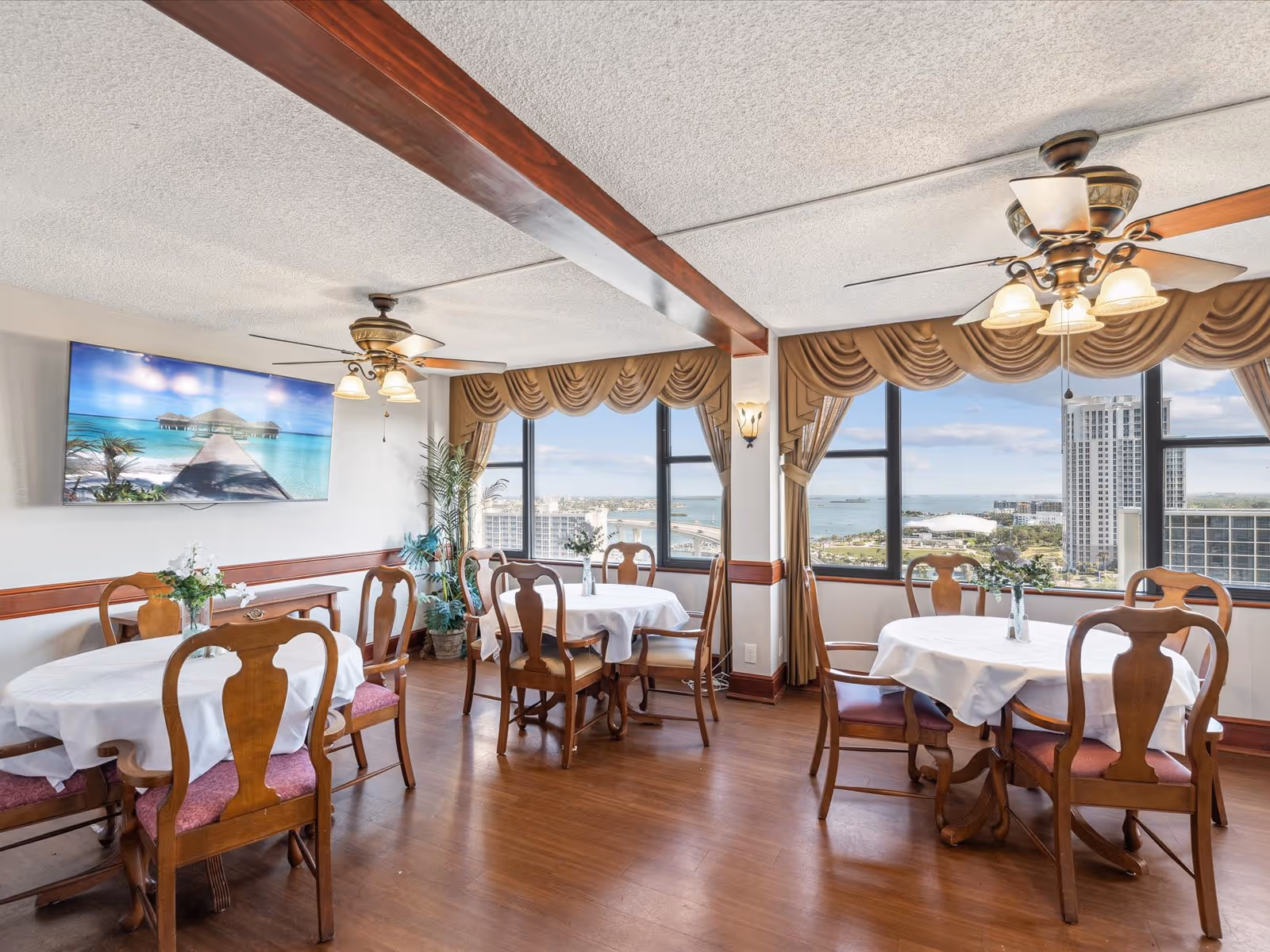 Dining room with round tables covered with white tablecloths and wooden chairs with red cushions. Large windows with brown drapes offer a view of a cityscape and water. Ceiling fans with lights hang from the ceiling, and a flat-screen TV displays an image of a pier over clear blue water. There are plants and floral centerpieces on the tables.