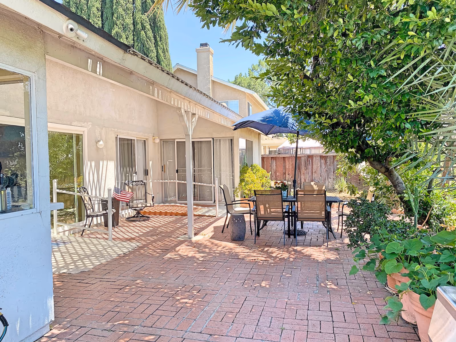 Outdoor patio area at Castlemere Senior Homes with brick flooring, a table with six chairs under a blue umbrella, potted plants, and a tree providing shade. The patio is adjacent to a building with sliding glass doors and windows.