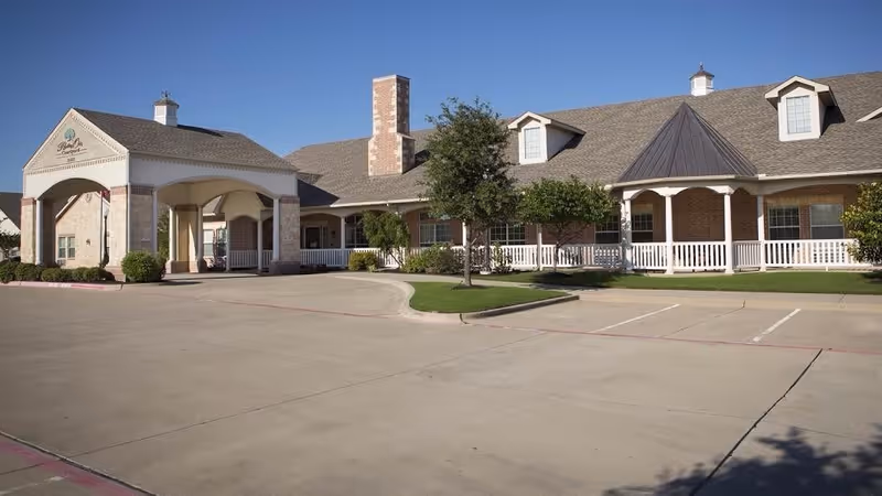 Exterior view of Bethesda Gardens Frisco Assisted Living facility showing a large brick building with a covered entrance, white railings, several windows, a chimney, and a parking area in front under a clear blue sky.