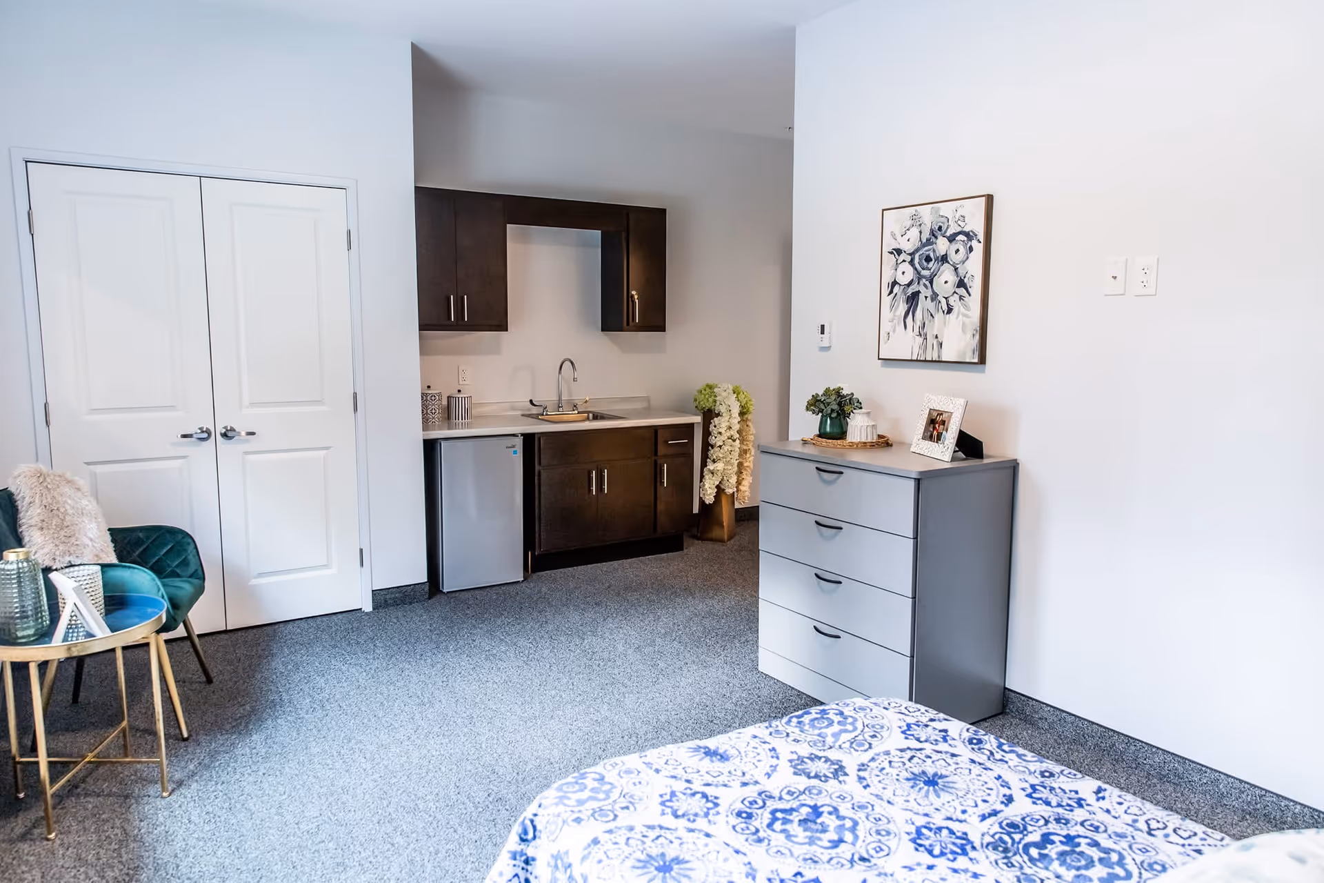 A bright and clean room featuring a small kitchenette with dark cabinets, a mini fridge, and a sink. There is a gray dresser with decorative items and a framed picture on top. A bed with a blue and white patterned bedspread is partially visible in the foreground. A green chair with a fur throw and a small round table with decorative items are placed near white double doors.
