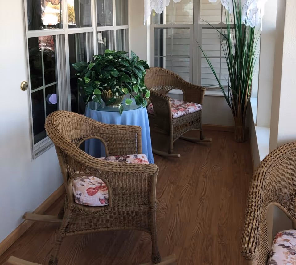 A cozy indoor seating area with three wicker rocking chairs featuring floral cushions, a small round table covered with a blue cloth and a green potted plant on top, wooden flooring, and large windows with white lace curtains letting in natural light.