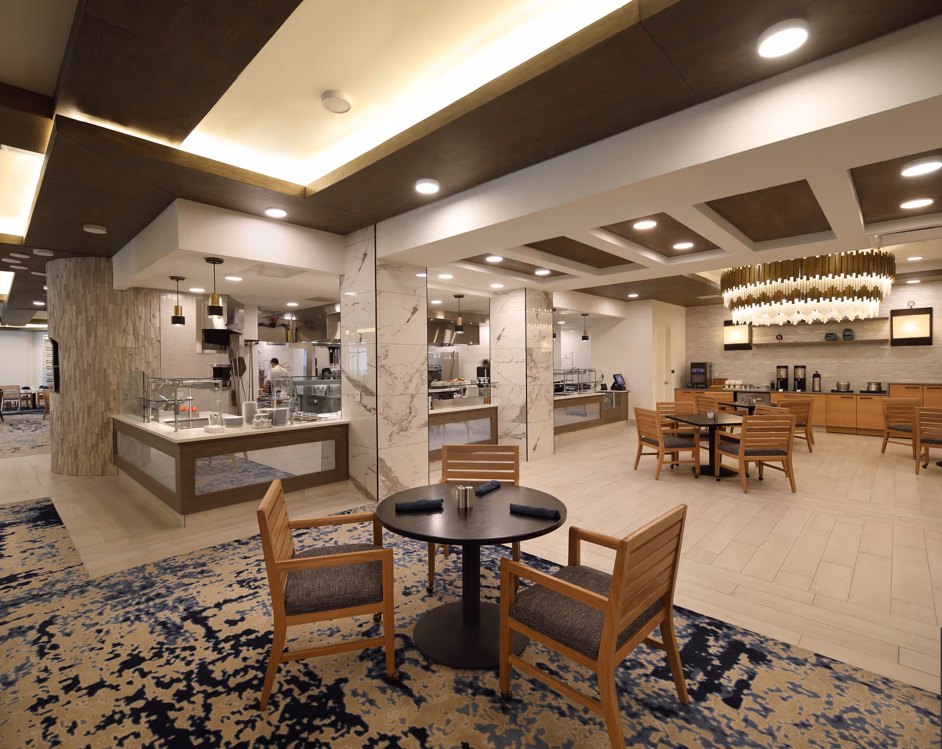 A modern dining area in a senior living facility with wooden chairs and round tables set with napkins and salt and pepper shakers. The space features a patterned carpet, marble pillars, and a buffet area with food service counters and coffee machines. The ceiling has recessed lighting and a large decorative chandelier.