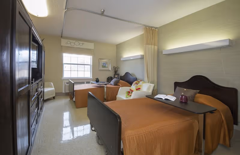 A shared bedroom in a senior living facility with two beds, a chair, and a window.