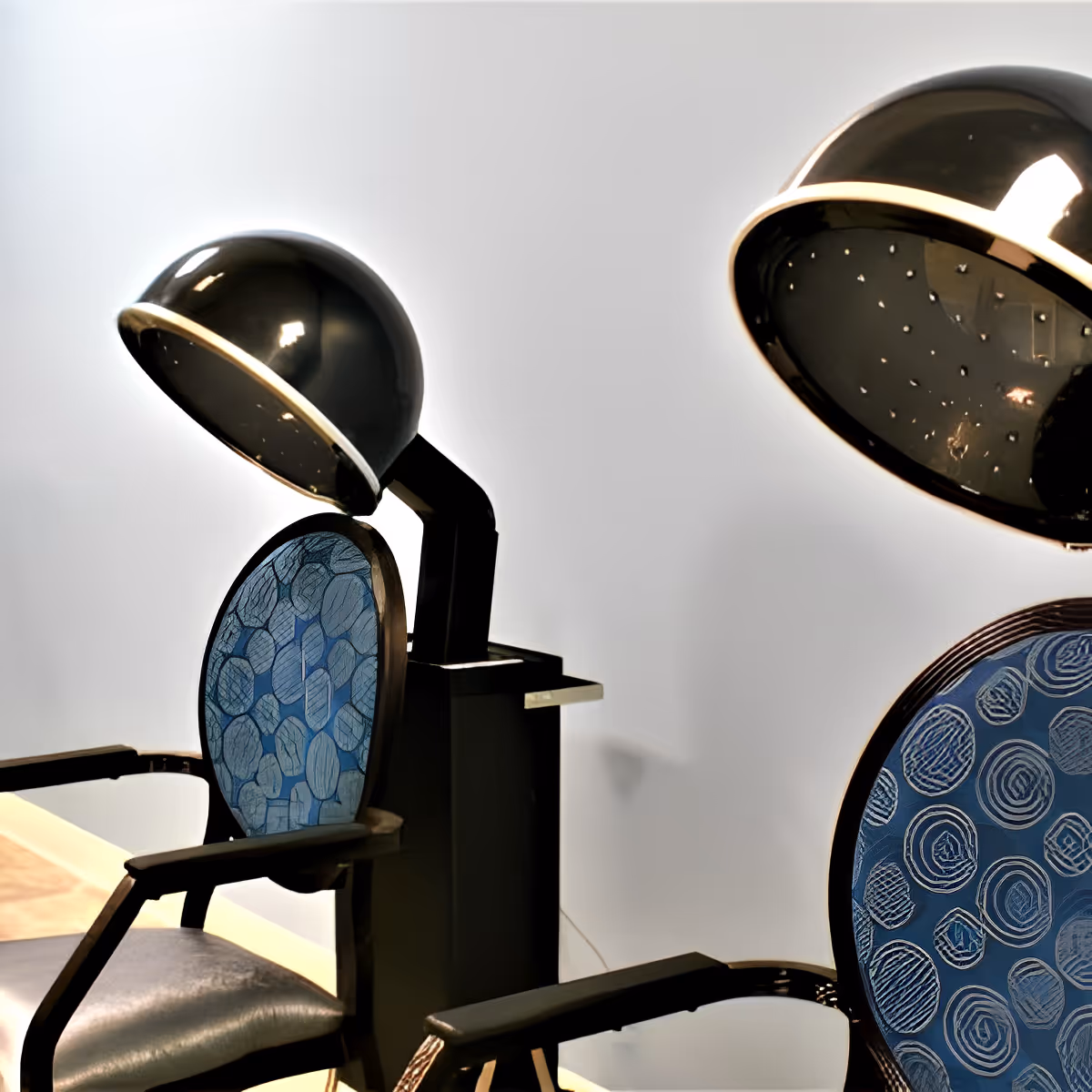 Two vintage-style hair drying chairs with blue patterned backs and black seats, each equipped with a black hooded hair dryer, set against a plain light-colored wall.