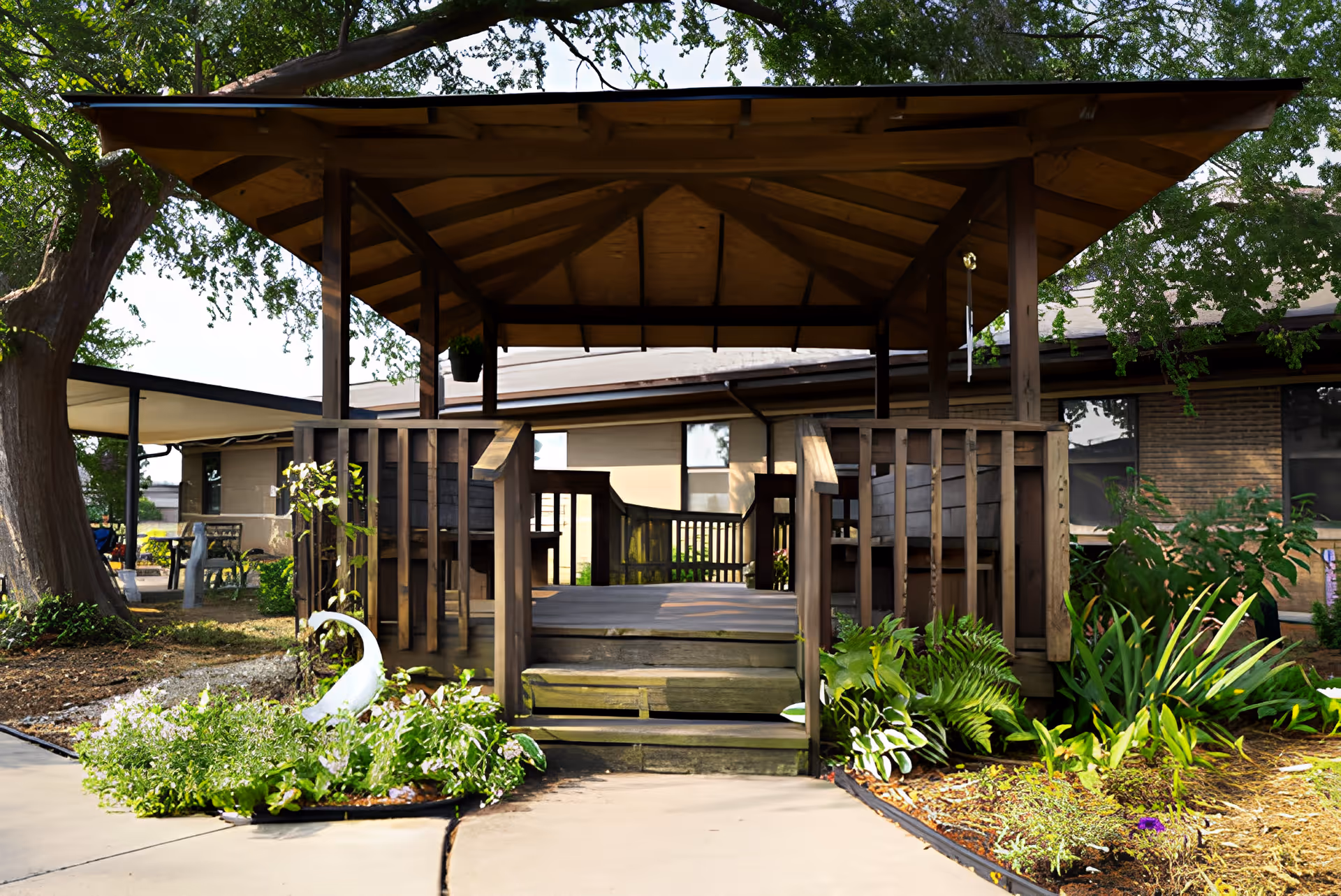 A wooden gazebo with a peaked roof and steps leading up to it, surrounded by green plants and flowers. The gazebo is located outdoors near a building with large windows and brick walls, with trees providing shade in the background.
