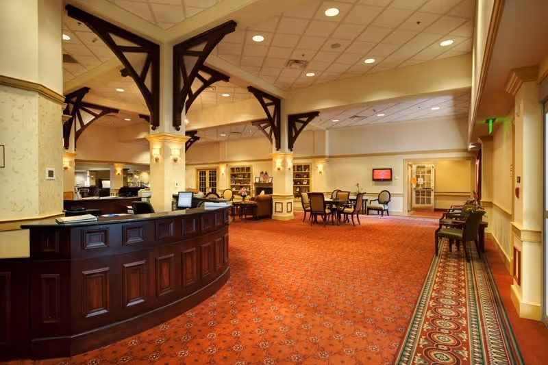 Spacious interior of a senior living facility with a large wooden reception desk on the left, red patterned carpet, several tables and chairs arranged for seating, decorative columns with dark wooden beams, and warm lighting. There is a hallway on the right side with additional chairs along the wall and a door at the end.