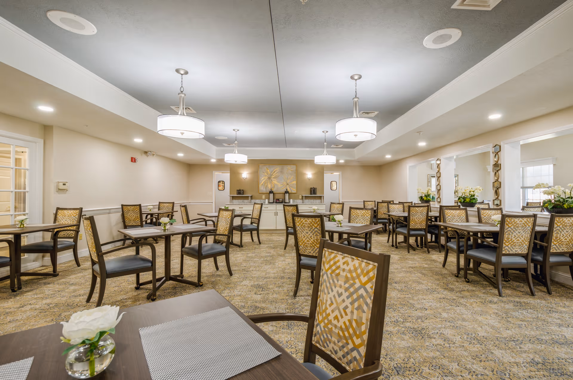 A spacious dining room with multiple tables and chairs arranged neatly. The room features beige walls, carpeted floors, and modern ceiling lights. There are decorative plants on some tables and a large artwork on the far wall above a sideboard with coffee dispensers.