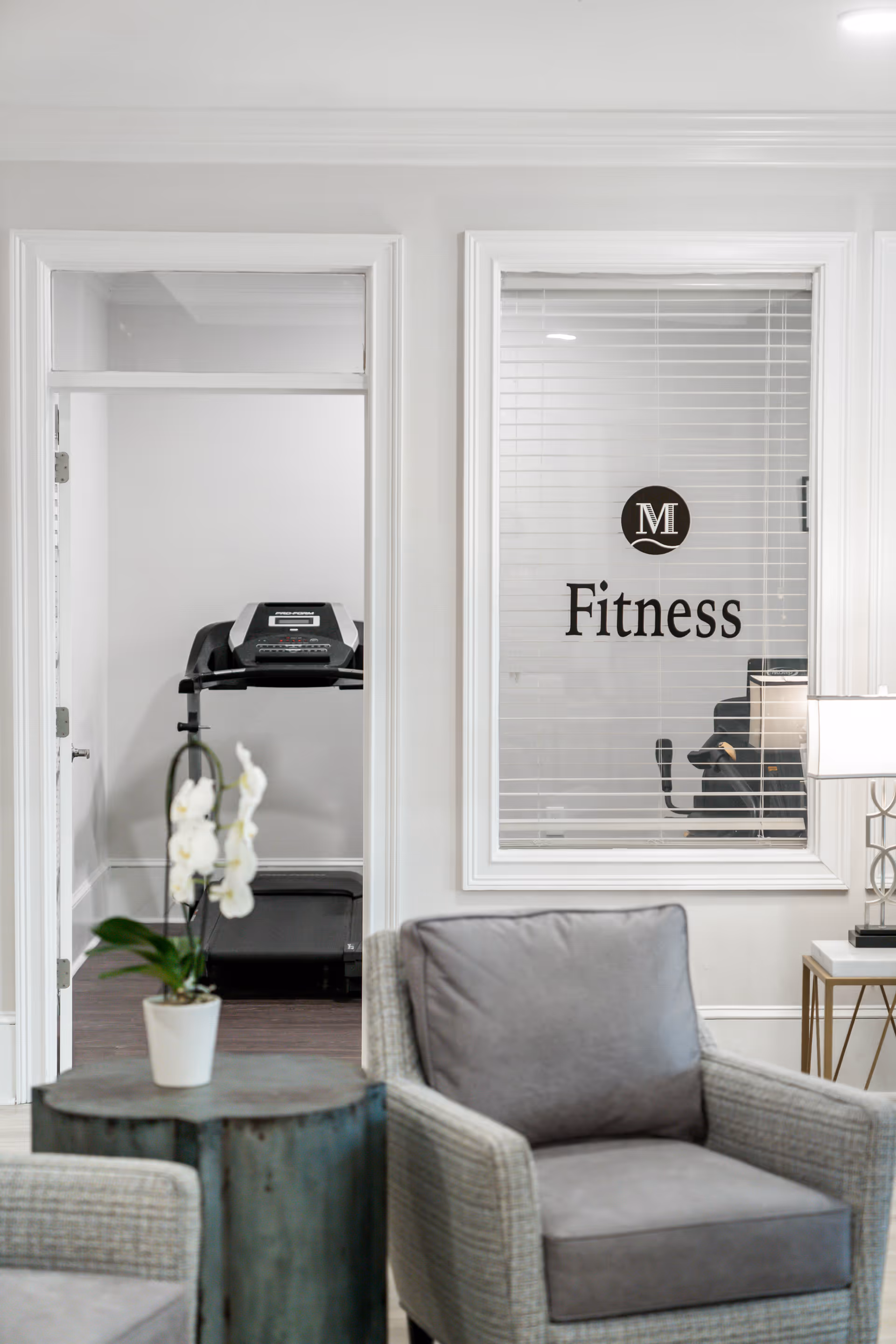 View of a fitness room through a glass window and open door from a seating area with two gray armchairs and a small round table with a white orchid plant. Inside the fitness room, a treadmill is visible. The glass window has the word 'Fitness' and a logo with the letter 'M'.