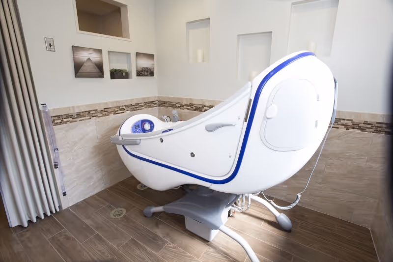 A modern, white and blue assisted bathing tub in a tiled bathroom with beige and brown tones. The tub has control buttons and handles, and the room features decorative wall niches with candles and framed pictures on the wall.
