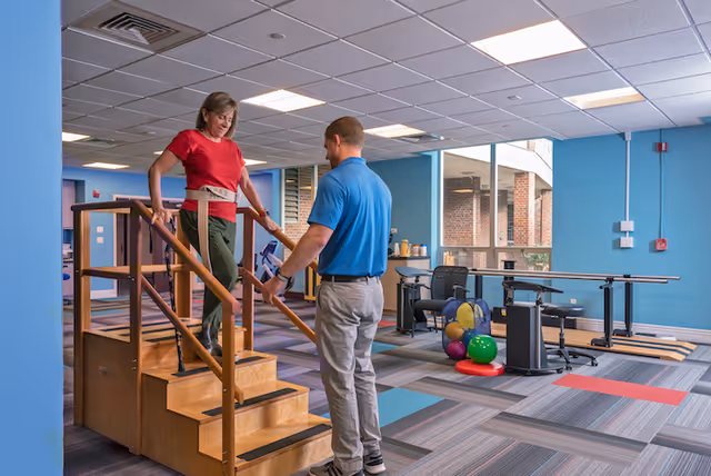 A woman wearing a red shirt and green pants is walking down a small wooden staircase with handrails inside a rehabilitation or therapy room. A man in a blue shirt and gray pants is assisting her. The room has blue walls, large windows, exercise equipment, and colorful therapy balls on the floor.
