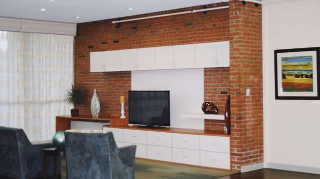 A cozy living room area featuring a brick accent wall with white cabinets and drawers. A flat-screen TV is placed on a wooden console, accompanied by decorative items including a large vase, a candle holder, and a sculpture. Two dark upholstered chairs face the TV, and a framed colorful landscape painting hangs on the adjacent white wall. Light-colored curtains cover a large window on the left side.