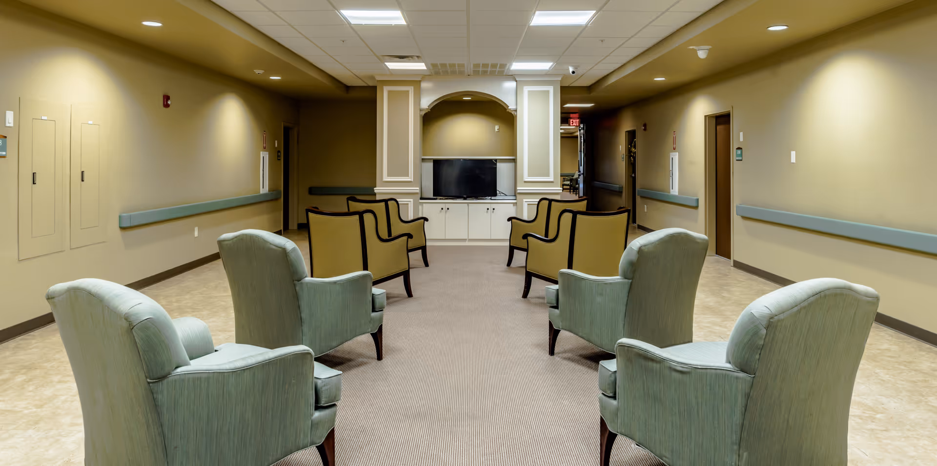 A seating area in a hallway of a senior living facility with eight armchairs arranged in two rows facing a TV mounted on a cabinet. The walls are beige with green handrails, and there are doors and utility panels along the hallway.
