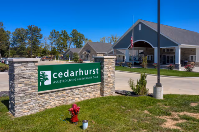 Exterior view of Cedarhurst assisted living and memory care facility in Lebanon, showing the main building with a covered entrance, an American flag, a stone sign with the Cedarhurst logo, and a parking area surrounded by trees under a clear blue sky.
