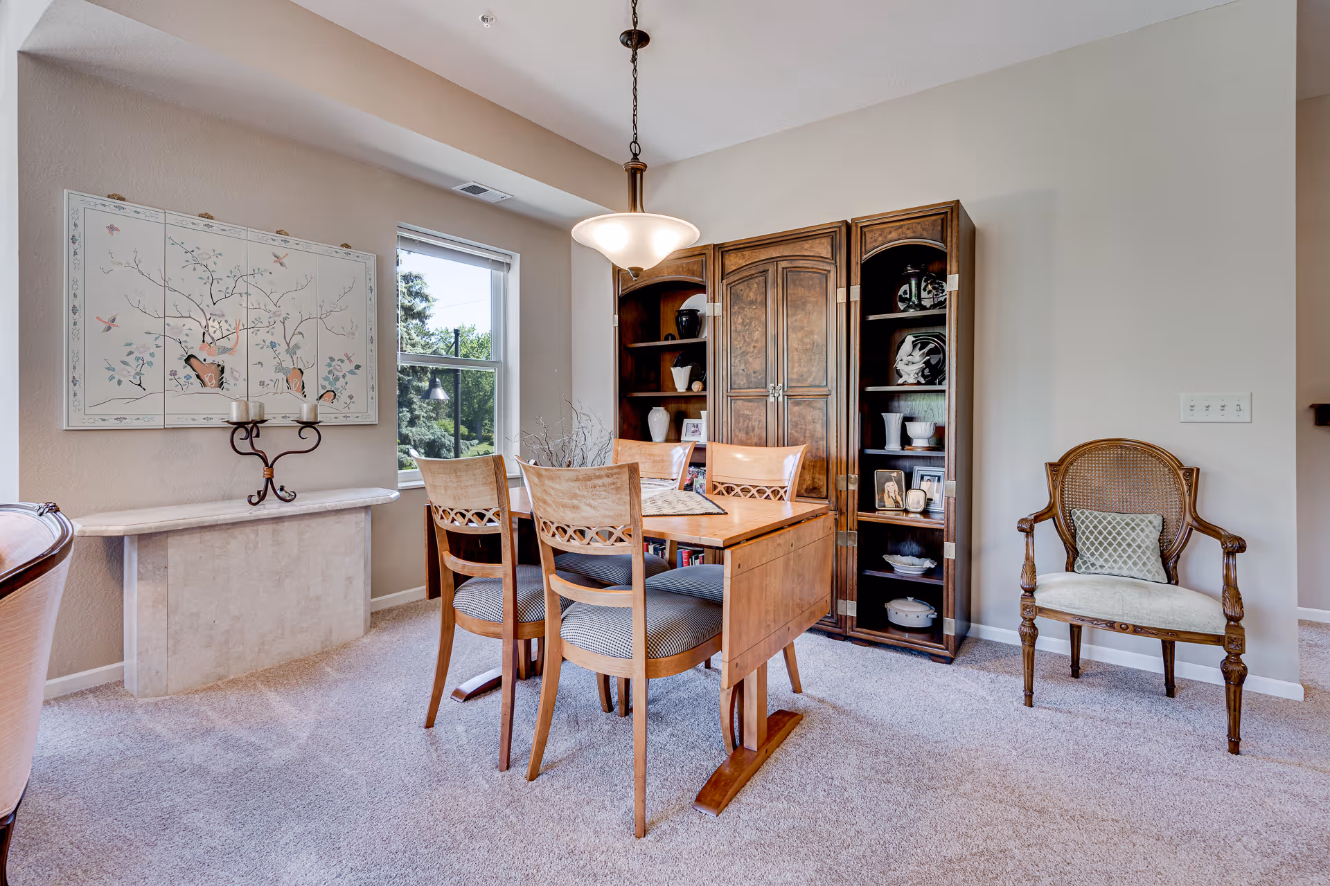 A dining room with a wooden table and four chairs, a wooden cabinet with shelves displaying decorative items, a single upholstered chair with a cushion, a window letting in natural light, and a wall-mounted artwork above a small marble console table.