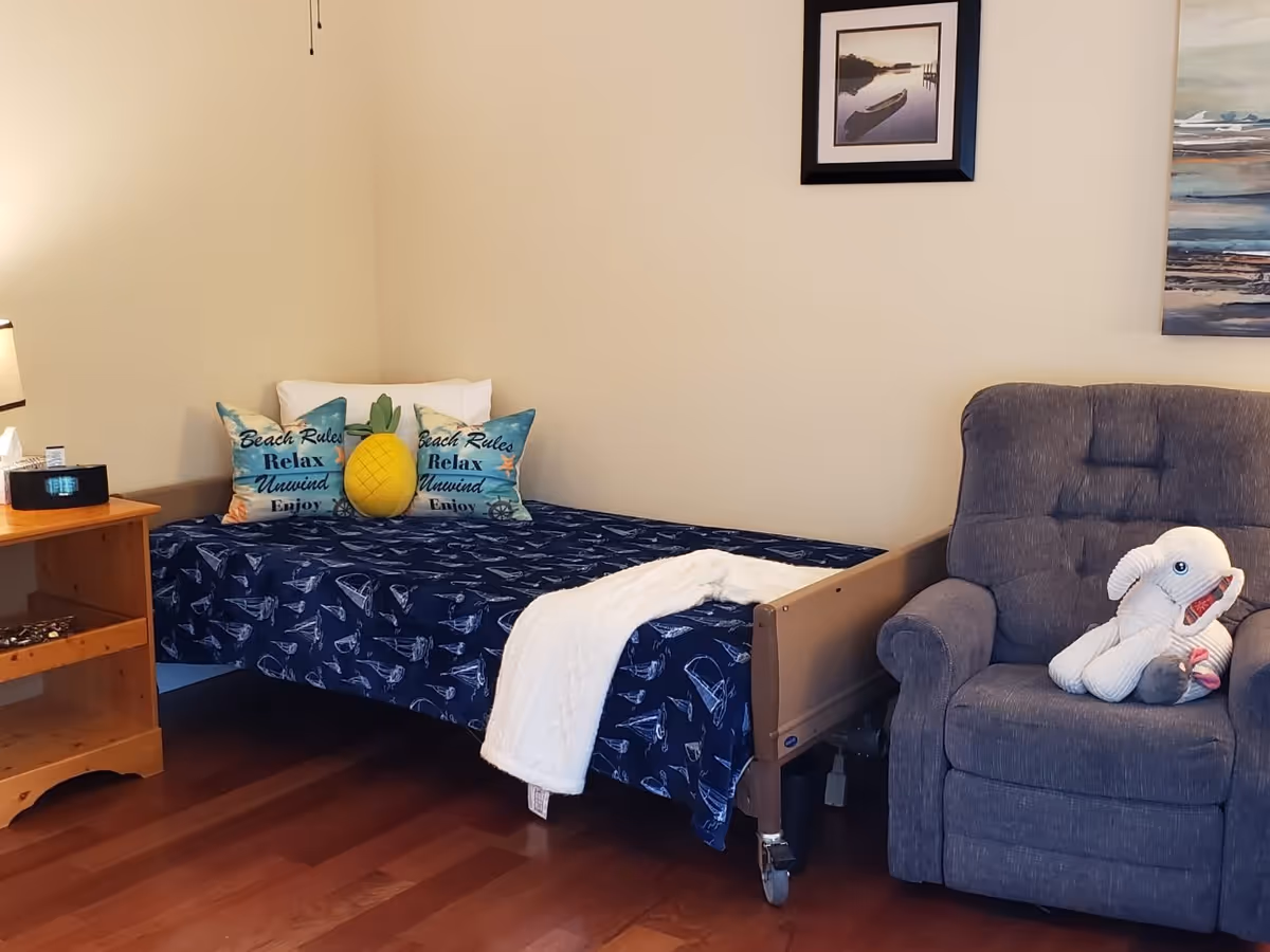 A cozy bedroom with a single bed covered in a blue bedspread featuring boat patterns. The bed has two decorative pillows with the words 'Beach Rules Relax Unwind Enjoy' and a pineapple-shaped pillow. A white blanket is draped over the bed. Next to the bed is a wooden nightstand with a lamp and a clock. To the right of the bed is a gray upholstered armchair with a stuffed white elephant toy on it. The walls are light-colored with two framed pictures hanging above the bed and chair.