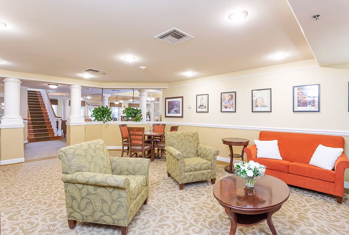 A bright and spacious senior living common area featuring two patterned armchairs, an orange sofa with white pillows, a round wooden coffee table with a flower arrangement, and a small side table. The room has beige walls adorned with framed photographs, carpeted flooring with a decorative pattern, and white columns separating the seating area from a dining space with a wooden table and chairs. A staircase is visible in the background.