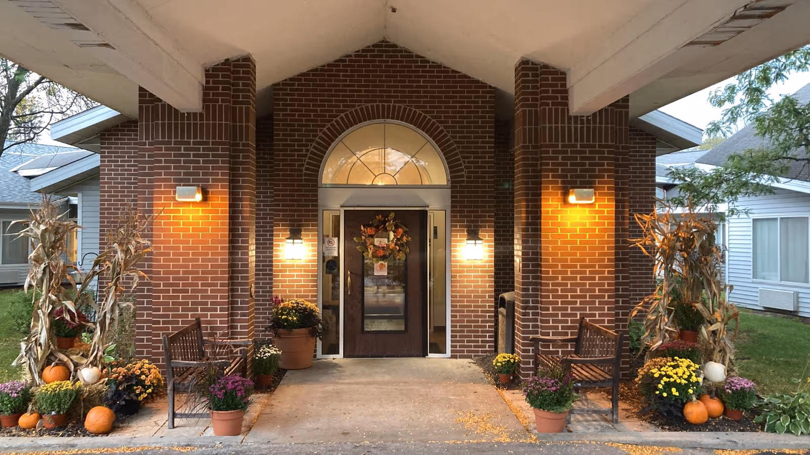 Entrance to a brick building with a covered porch, decorated with autumn-themed plants, pumpkins, and flowers. Two wooden benches are placed on either side of the entrance door, which has a decorative wreath. The area is lit by wall-mounted lights on the brick columns.