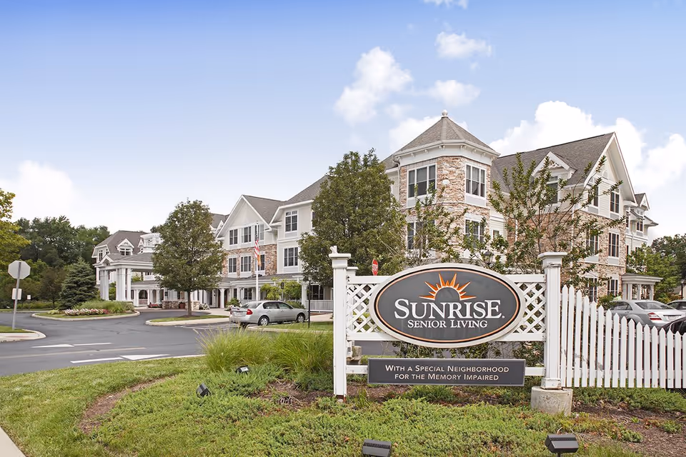 Exterior view of Sunrise Senior Living facility with a large sign in front that reads 'Sunrise Senior Living' and 'With a Special Neighborhood for the Memory Impaired'. The building is multi-story with white siding and stone accents, surrounded by trees and parked cars under a partly cloudy sky.
