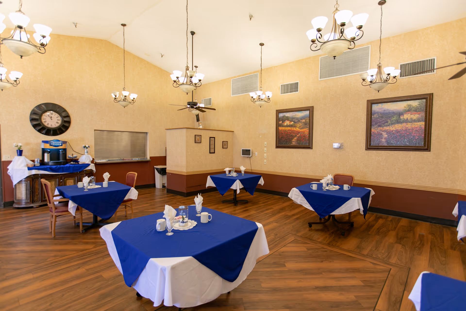 A dining room with several tables covered in white tablecloths and blue overlays, each set with cups, napkins, and small centerpieces. The room has wooden flooring, beige walls with two framed landscape paintings, multiple chandeliers hanging from the ceiling, and a large clock on one wall above a coffee station.