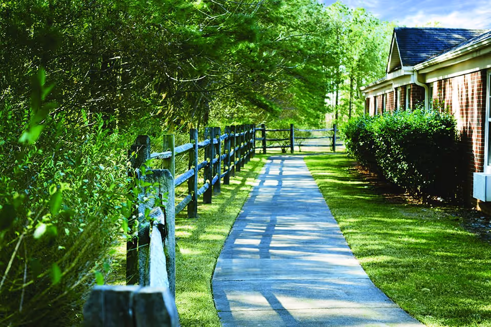 A paved walkway bordered by a wooden fence and green bushes on one side and a brick building with shrubs on the other side, under a bright blue sky with some clouds.
