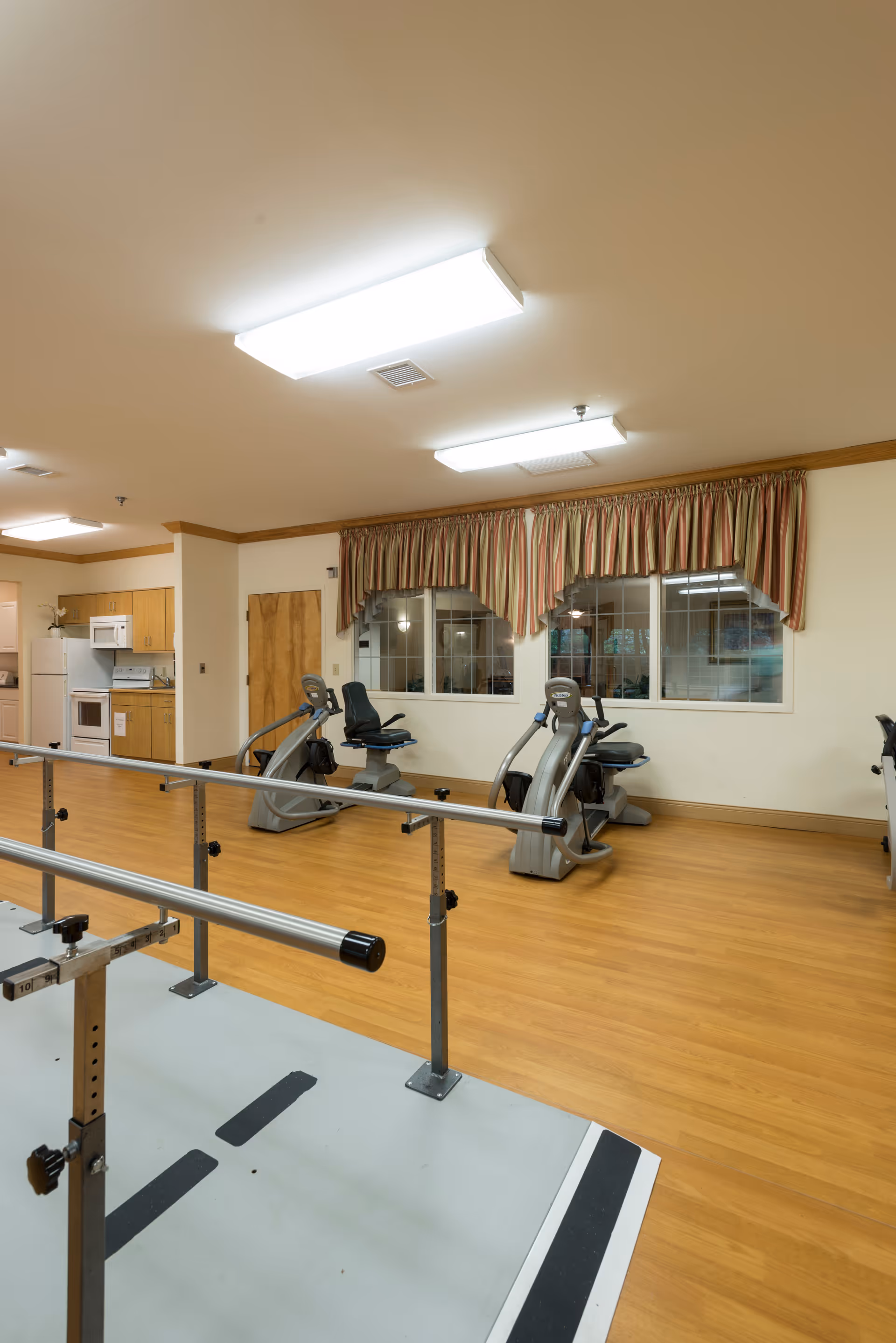 Interior view of a rehabilitation or exercise room in a skilled nursing facility with wooden flooring, parallel bars for walking support, three stationary exercise bikes, a kitchenette area with a refrigerator, microwave, and cabinets, and windows with striped curtains.