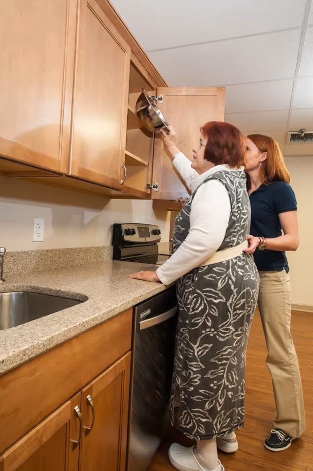 An elderly woman is reaching up to place or retrieve a metal bowl from a wooden kitchen cabinet while a caregiver stands behind her, providing support. The kitchen has wooden cabinets, a granite countertop, a stainless steel sink, and a stove.