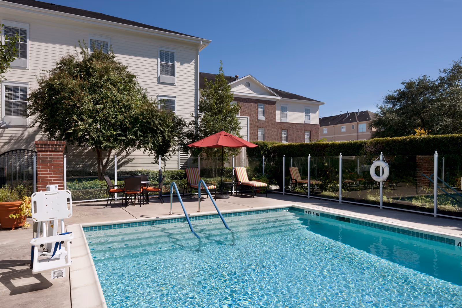 Outdoor swimming pool area at Belmont Village Senior Living West University with clear blue water, poolside chairs with cushions, a red umbrella, trees, and residential buildings in the background under a clear blue sky.