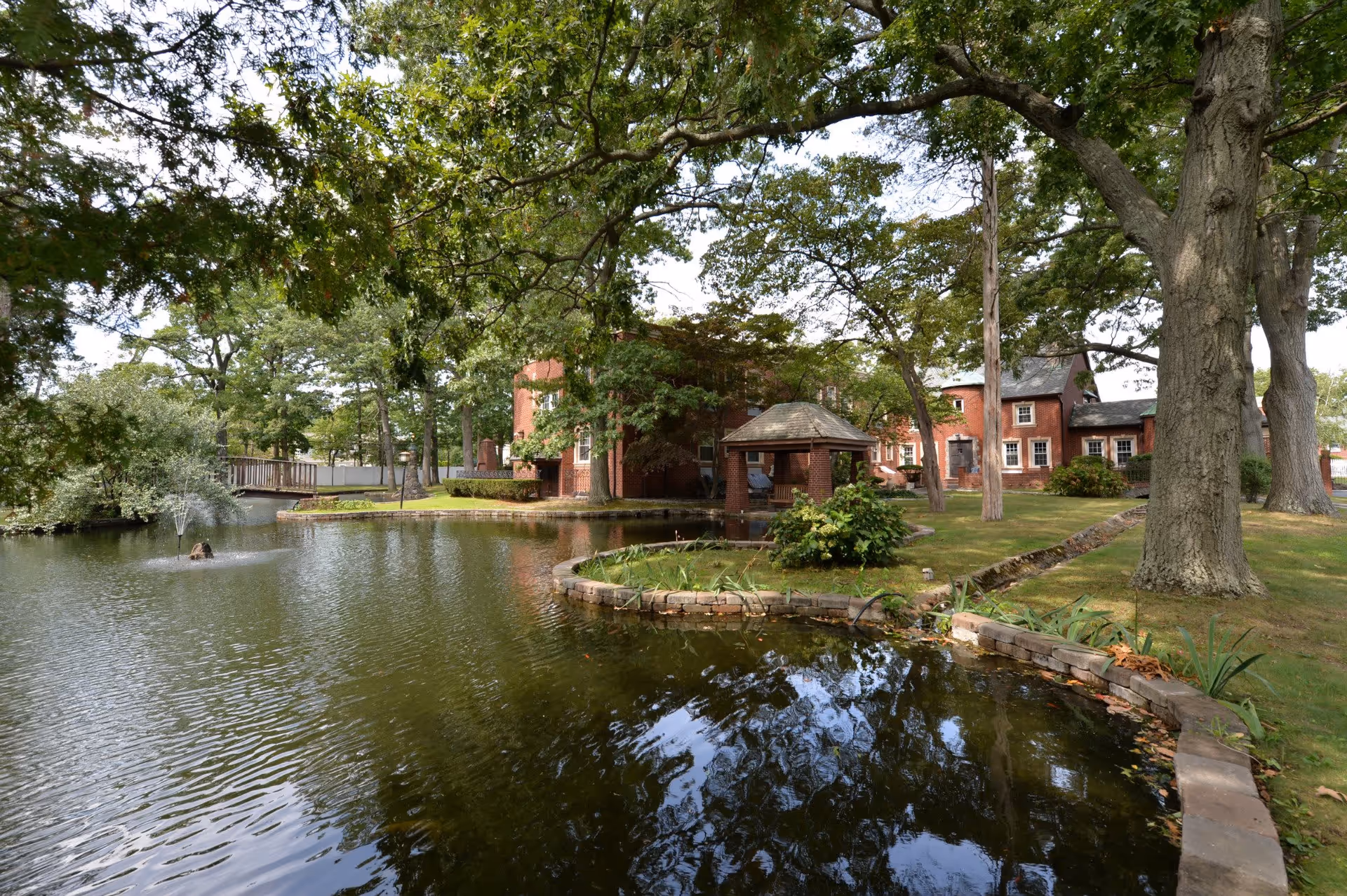 A serene outdoor scene at Birchwood Suites featuring a pond with a small fountain, surrounded by lush green trees and grass. There is a wooden bridge over the pond and a brick gazebo near the water's edge. In the background, a large red brick building is partially visible through the trees.
