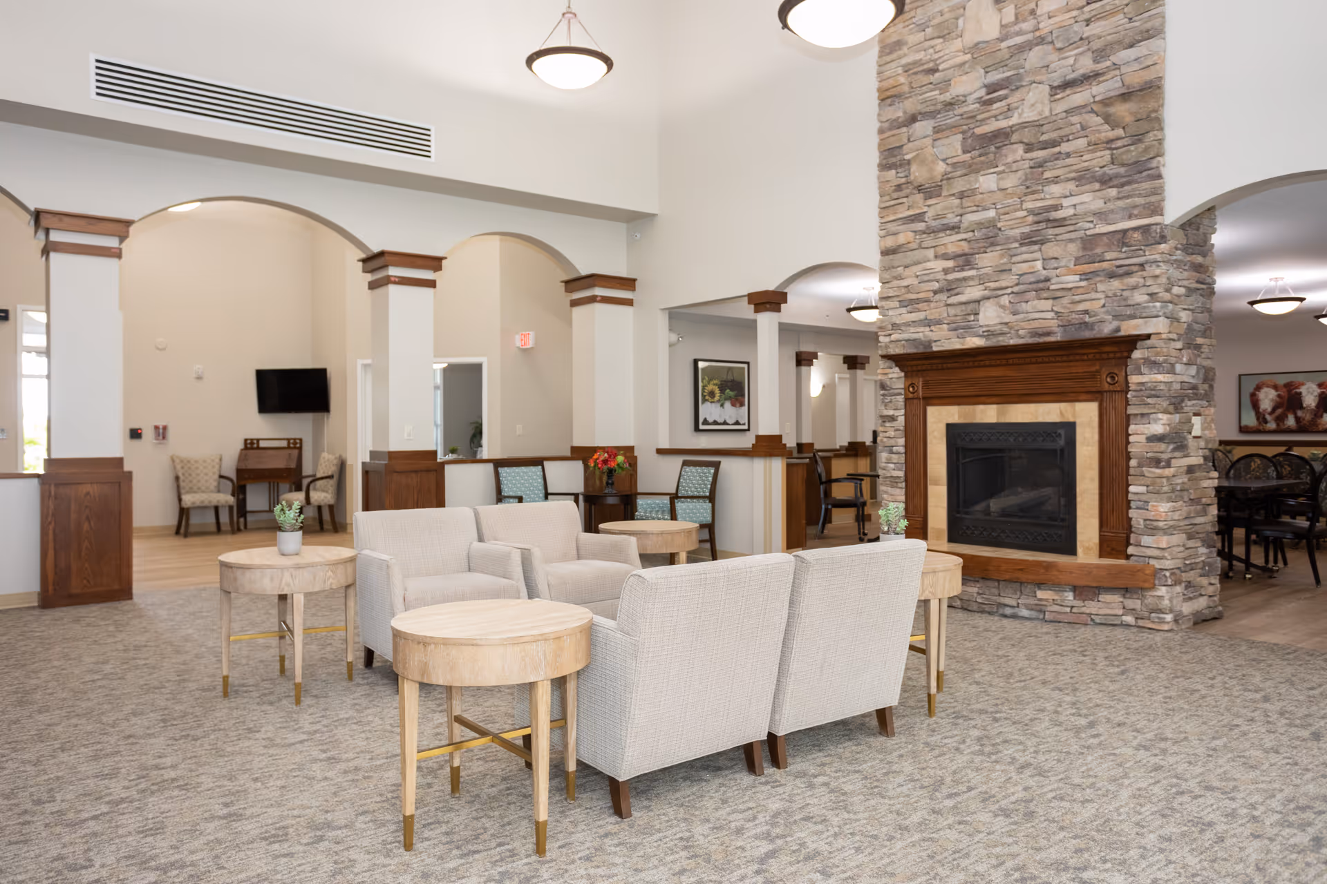 A spacious and well-lit living room area in a senior living facility featuring a stone fireplace with a wooden mantle, beige upholstered armchairs arranged around light wood round tables, and archways leading to other rooms. The room has carpeted flooring, neutral-colored walls, and ceiling lights. In the background, there are additional seating areas and a wall-mounted TV.