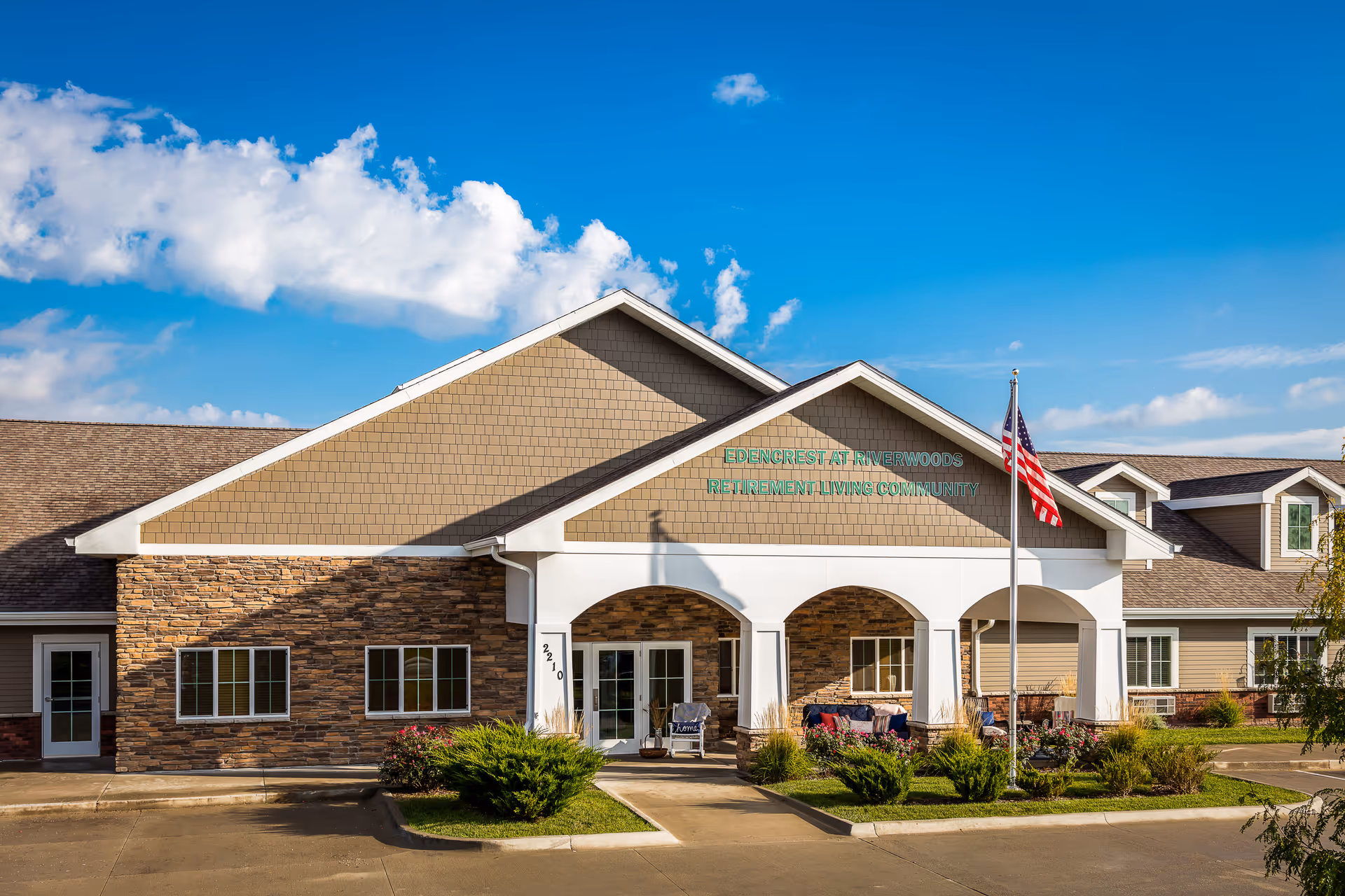 Front exterior view of Edencrest at Riverwoods retirement living community building with a clear blue sky, American flag on a flagpole, and landscaped bushes and flowers near the entrance.