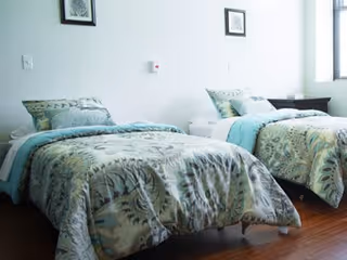 A bright, clean bedroom with two twin beds featuring matching patterned bedspreads and pillows. The room has wooden flooring, a small dark wooden nightstand between the beds, and framed artwork on the white walls.