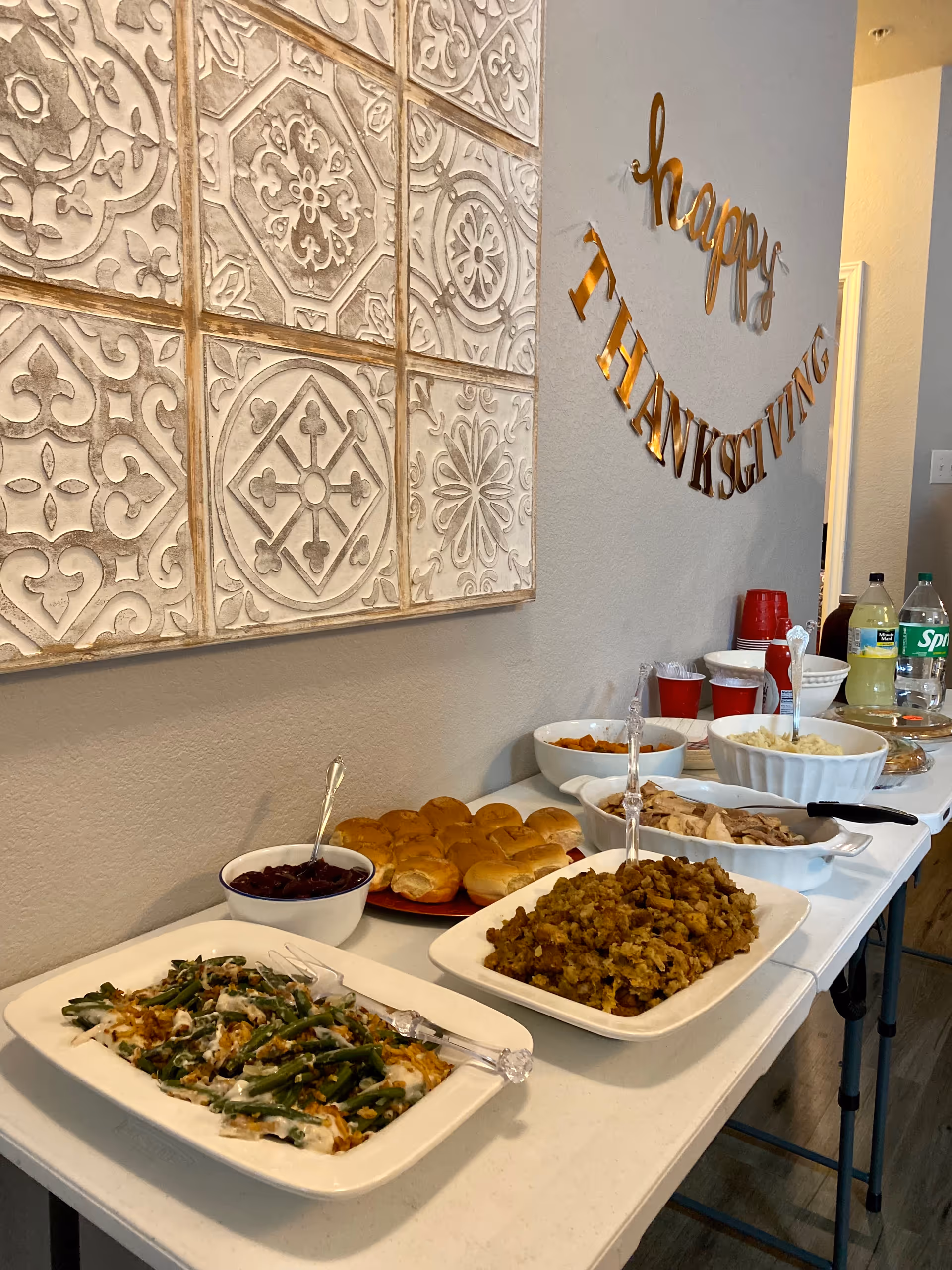 A buffet table set up with various Thanksgiving dishes including green bean casserole, stuffing, dinner rolls, cranberry sauce, sliced turkey, mashed potatoes, and beverages. A decorative wall hanging reads 'happy THANKSGIVING' in gold letters above the table.