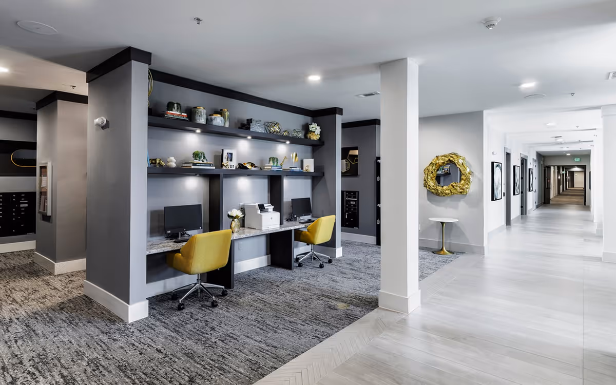 Bright interior hallway with a modern communal work area featuring two yellow chairs, computers and shelving on the left.