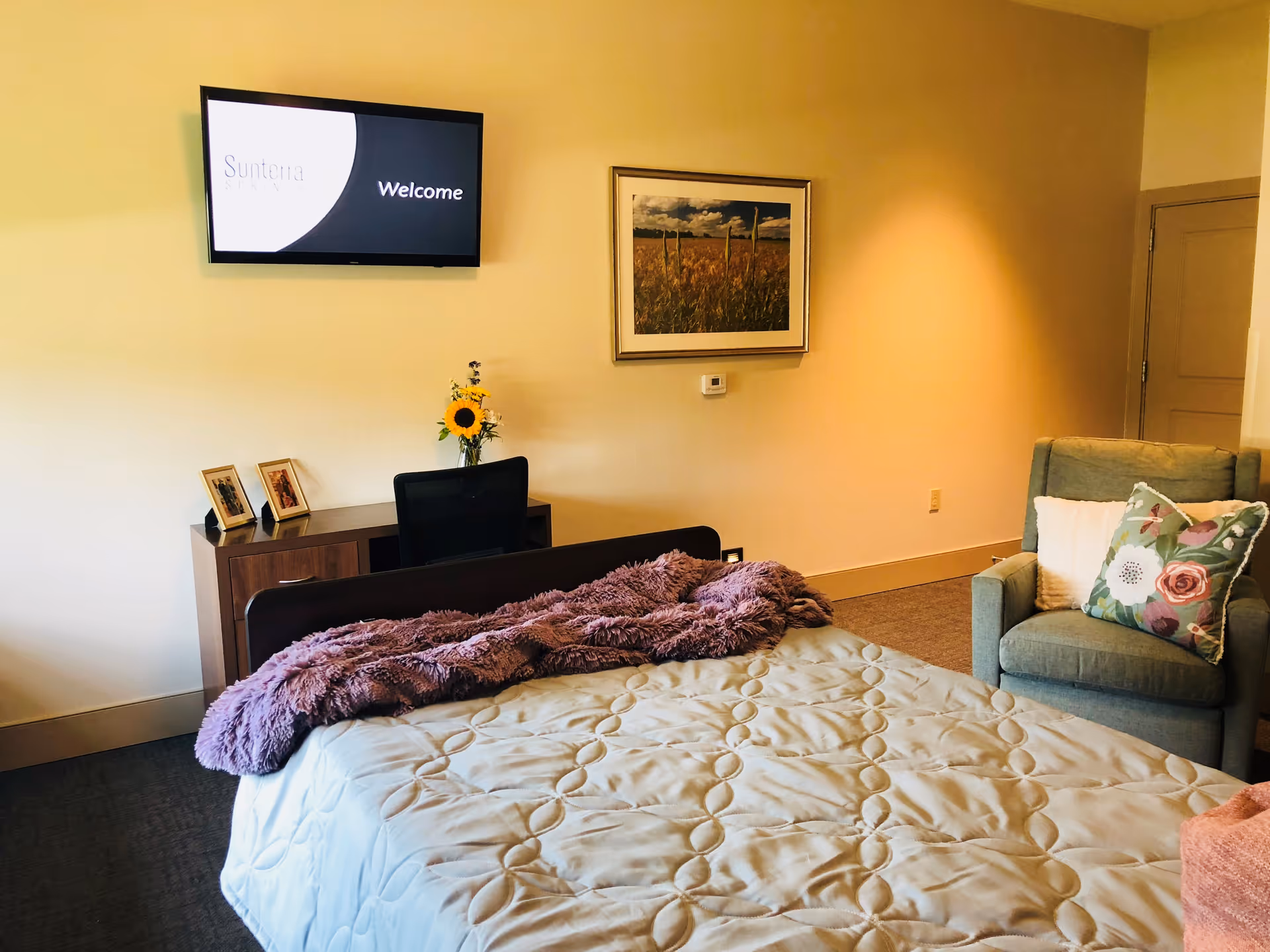 A cozy bedroom in a senior living facility featuring a bed with a white quilted comforter and a purple throw blanket. There is a green armchair with floral and white pillows to the right. A wooden desk with a black office chair, two framed photos, and a vase with a sunflower is against the wall. Above the desk, a wall-mounted TV screen displays the text 'Sunterra Welcome'. A framed picture of a field with a blue sky hangs on the wall.