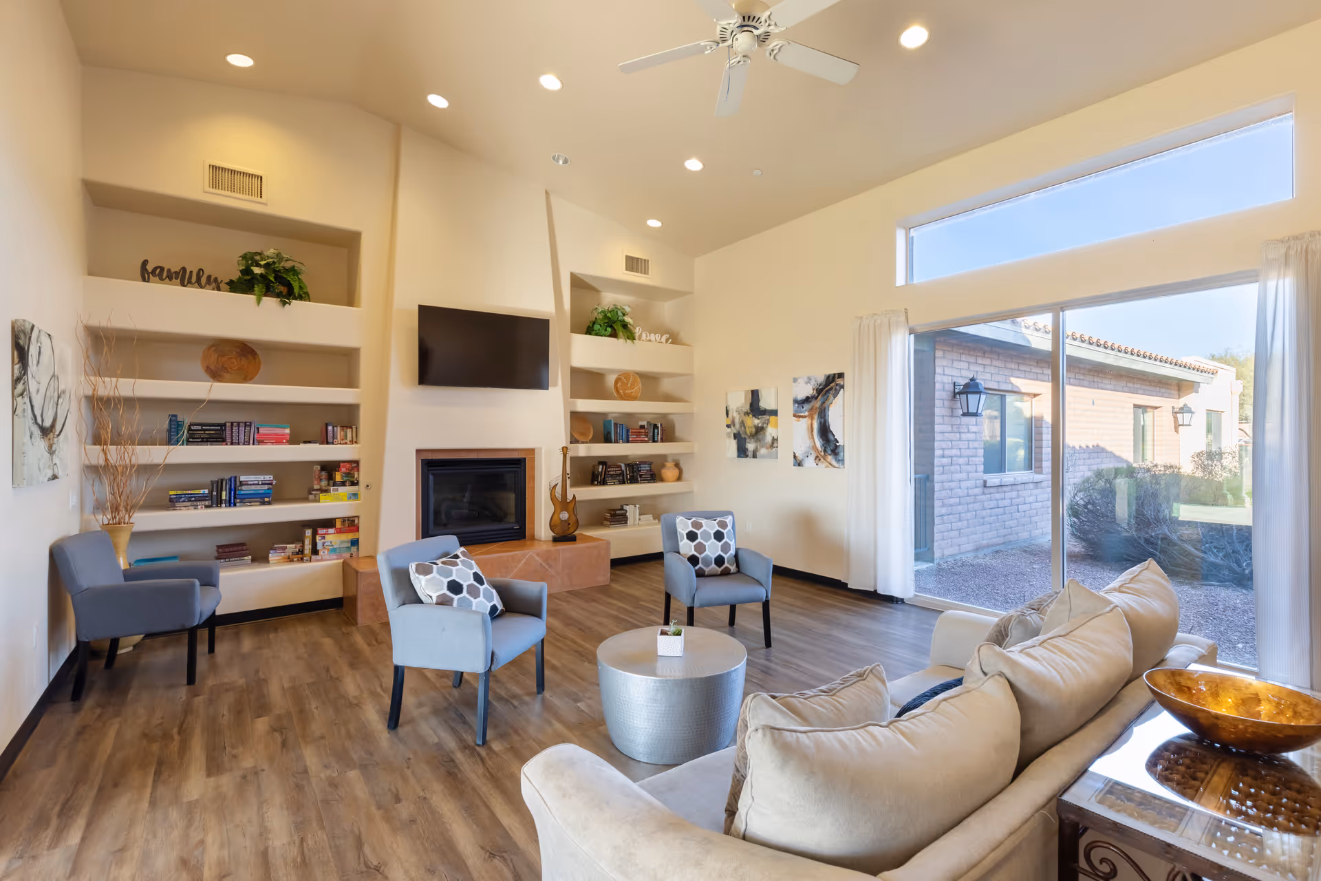 A bright and spacious living room with wooden flooring, beige walls, and a high ceiling with recessed lighting and a ceiling fan. The room features a beige sofa with cushions, two blue armchairs with patterned pillows, a round silver coffee table, built-in white shelves with books and decorative items, a wall-mounted TV above a fireplace, and large windows with white curtains letting in natural light and showing an outdoor view of a brick building and garden.