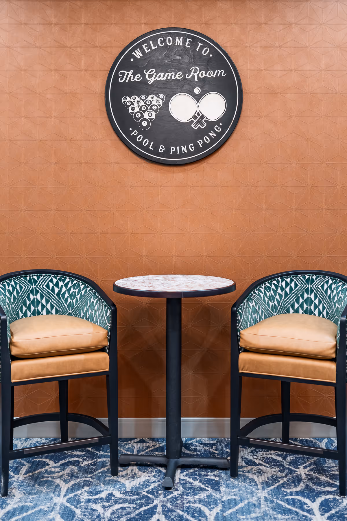 Two cushioned chairs with patterned backs and tan seats are positioned on either side of a small round table with a marble top, set against a textured brown wall. Above the table is a circular sign that reads 'Welcome to The Game Room Pool & Ping Pong' with illustrations of billiard balls and ping pong paddles.