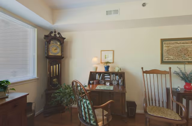A cozy interior room featuring a wooden grandfather clock, a wooden desk with a lamp, plants, and decorative items, two wooden chairs with cushions, and framed artwork on the walls.