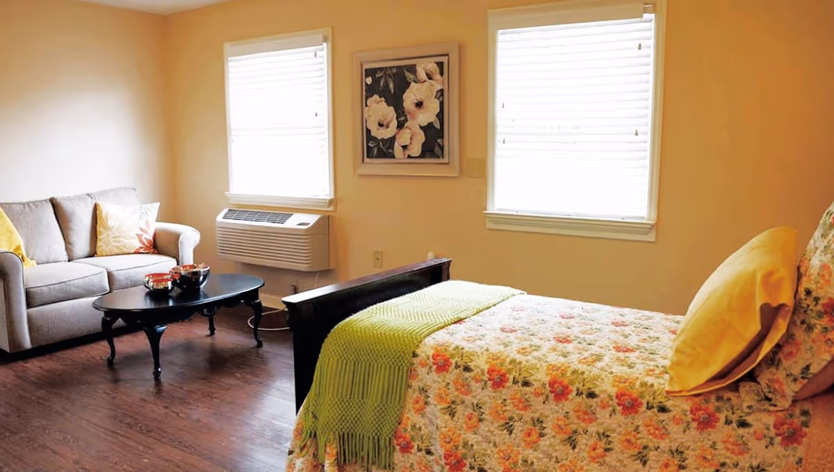 A cozy senior living room and bedroom area with a floral bedspread and yellow pillows on the bed, a beige sofa with decorative pillows, a black coffee table, two windows with white blinds, and a floral painting on the wall.