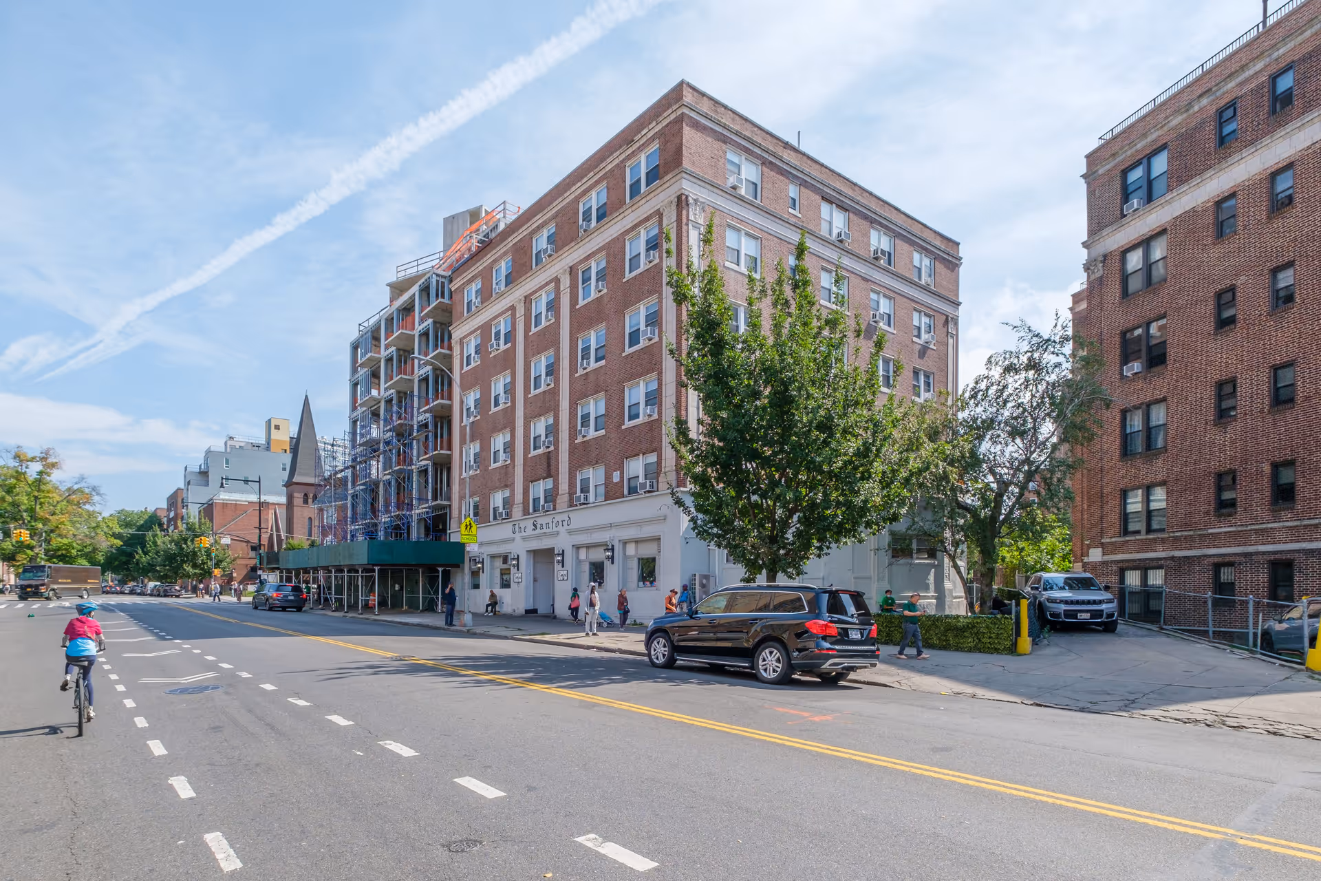 Street view of a multi-story brick building named The Sanford with people walking on the sidewalk and cars parked along the street. There is a cyclist riding on the road and trees lining the sidewalk under a partly cloudy sky.