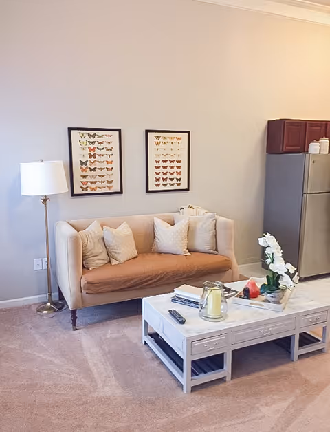 A cozy living area featuring a beige loveseat with four decorative pillows, a white coffee table with a candle, remote control, books, and a flower arrangement. A floor lamp stands to the left of the loveseat, and a stainless steel refrigerator with wooden cabinets is visible on the right. Two framed butterfly prints hang on the wall above the loveseat.