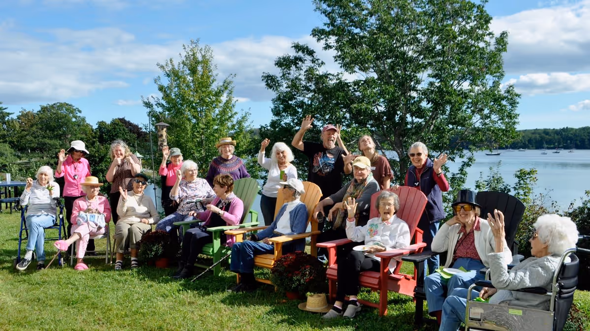 A group of elderly people sitting outdoors on colorful Adirondack chairs and lawn chairs, smiling and waving at the camera. They are on a grassy area near a body of water with trees and a partly cloudy sky in the background.