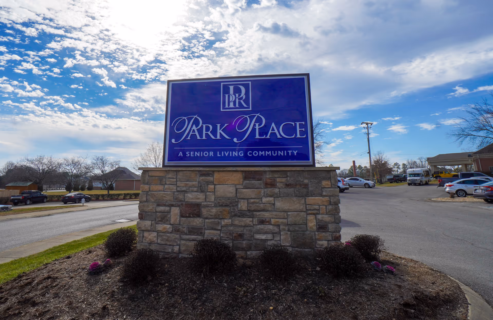 A large stone sign for Park Place, a senior living community, stands in a landscaped area with bushes and mulch. The sky is partly cloudy with blue patches, and there are cars parked in the background along a paved road.