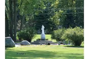 White statue of a robed figure on a pedestal in a grassy, tree-lined garden.