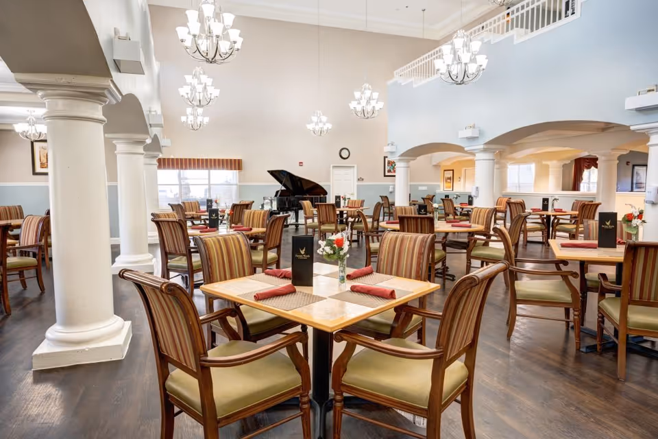 A spacious dining room with multiple wooden tables and chairs arranged neatly. Each table has a small vase with flowers and folded red napkins. The room features high ceilings with several chandeliers, white columns, and a grand piano near the back wall. The walls are painted in light colors, and there are large windows letting in natural light.