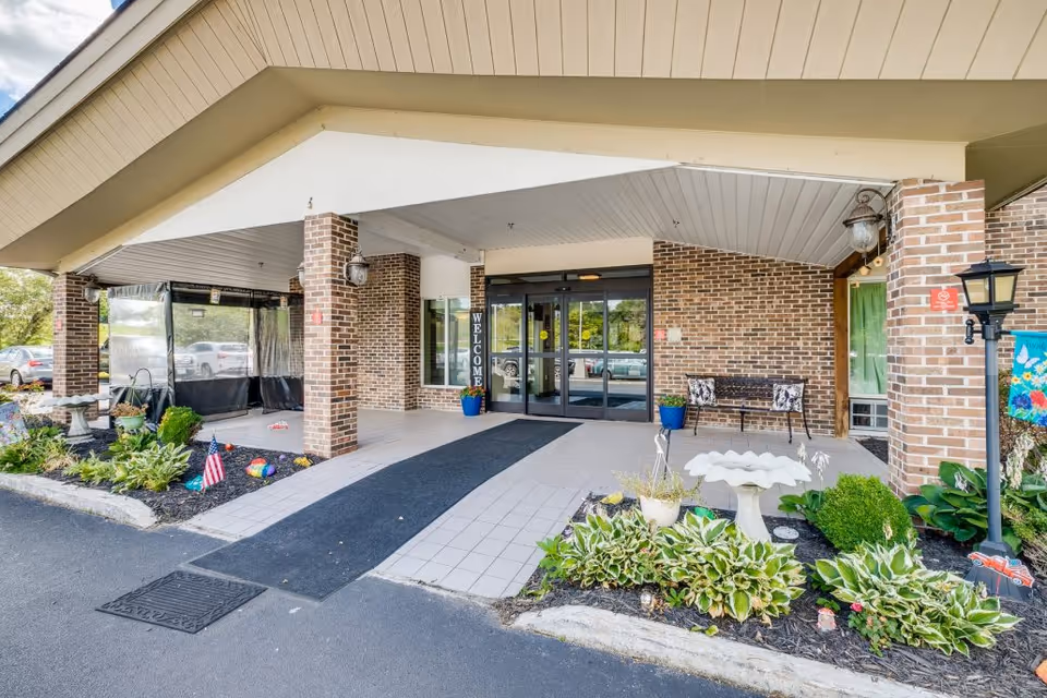 Entrance of Greystone Healthcare Center showing a covered driveway with brick pillars, a welcome sign near the glass doors, a bench, decorative plants, a birdbath, and a lamp post.