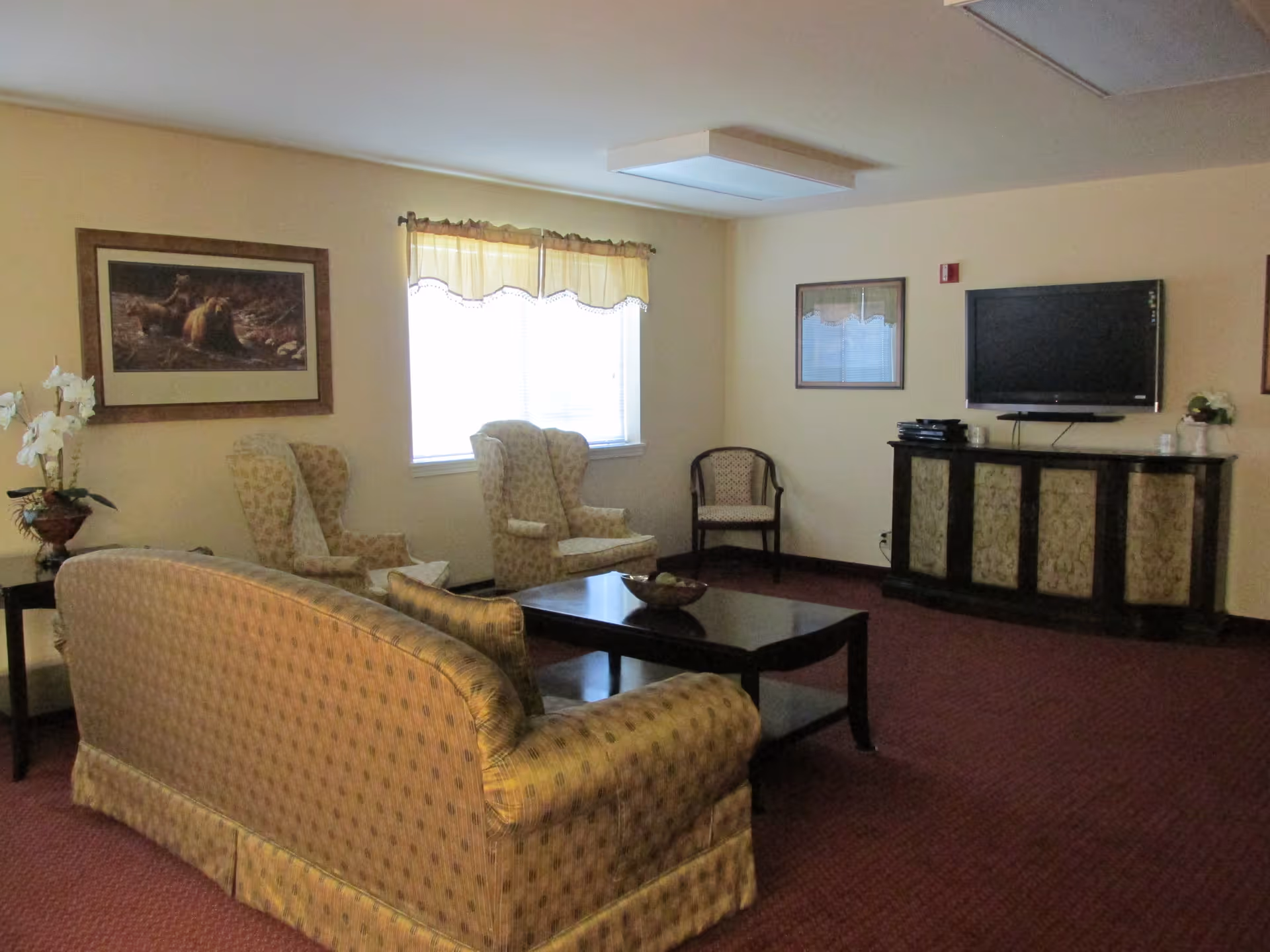 A cozy living room with patterned upholstered furniture including a sofa and two armchairs arranged around a dark wood coffee table. A flat-screen TV is mounted on the wall above a decorative cabinet. The room has beige walls, a window with a yellow valance, framed artwork, and a red carpeted floor.