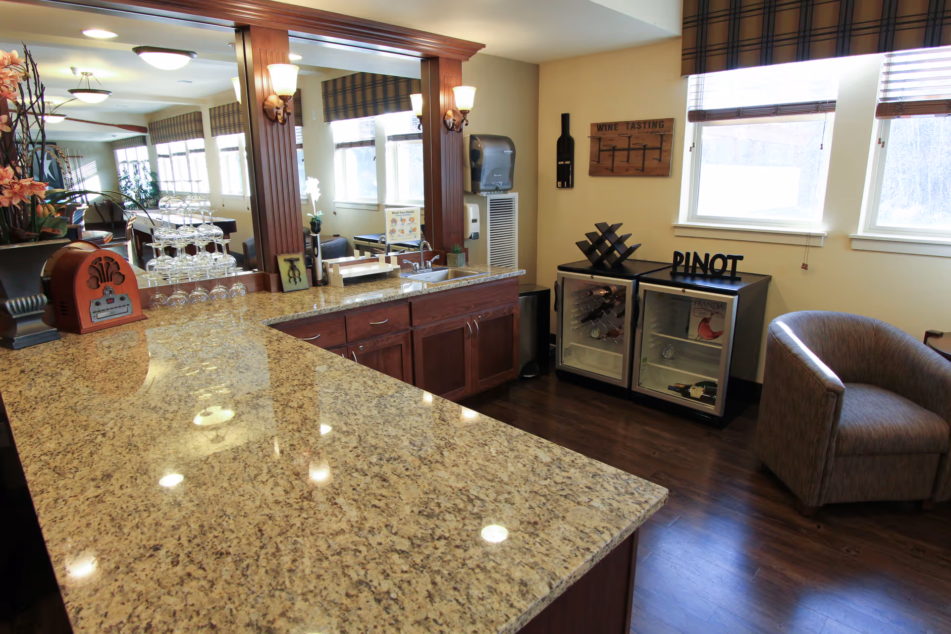 Interior view of a lounge area with a granite countertop bar, wooden cabinets, a large mirror with wall sconces, a small sink, and two mini refrigerators labeled with wine bottles. There is a comfortable armchair near the windows with blinds, and decorative items including a wine tasting sign and a wine rack.