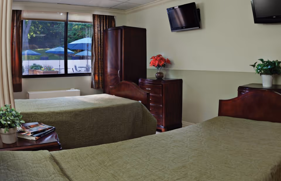 A bedroom with two twin beds covered in green quilts, a wooden nightstand with books and a plant, a wooden dresser with a vase of red flowers, a wooden wardrobe, a wall-mounted TV, and a window showing an outdoor area with blue umbrellas.