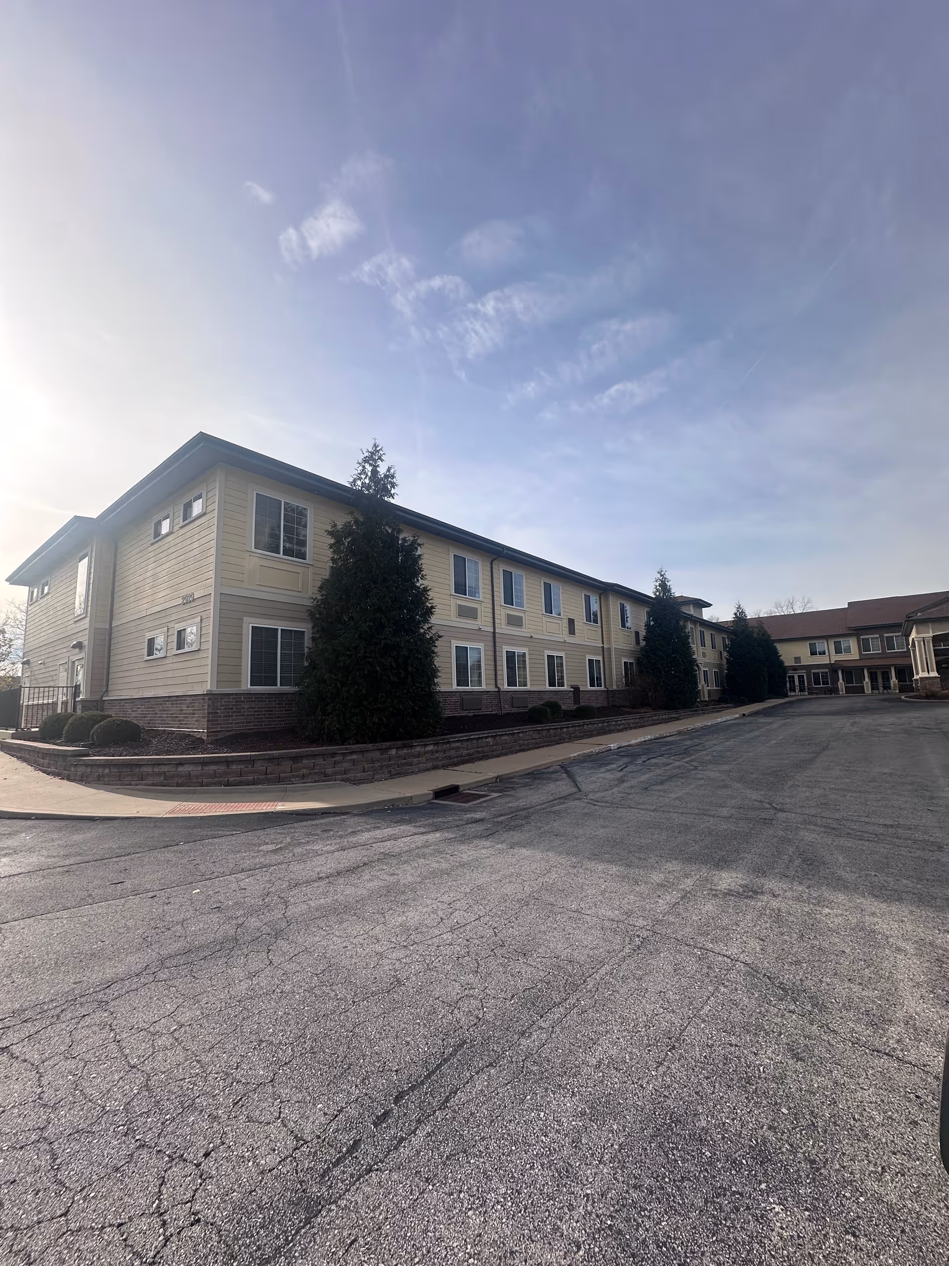 Exterior view of a two-story assisted living facility building with beige siding and brick accents, surrounded by a paved driveway and landscaped with evergreen trees and shrubs under a partly cloudy sky.