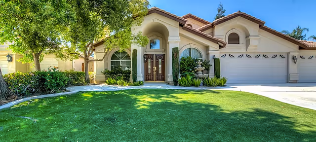 Front exterior of a single-story beige stucco building with an arched entryway, double wooden doors, attached three-car garage, fountain and a green lawn.